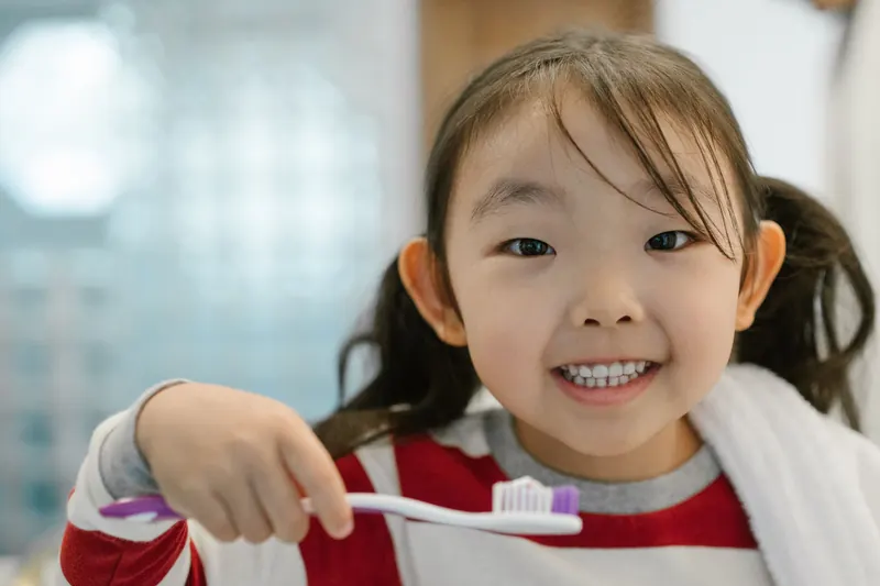 A smiling little girl holding up her toothbrush in a bright bathroom